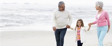 Una pareja de adultos mayores sonriendo, caminando por la playa cogidos de la mano de una niña pequeña.