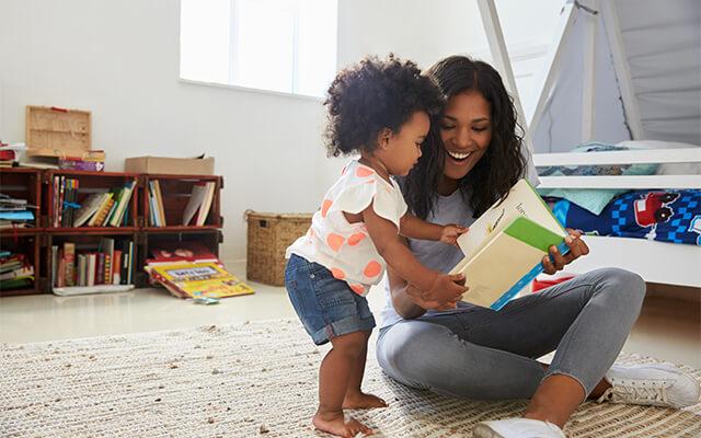 Una mama sentada en el suelo con su hija en una habitación infantil leyendo un cuento.